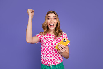 Smiling young woman with smartphone celebrates success with fist raised against colorful background