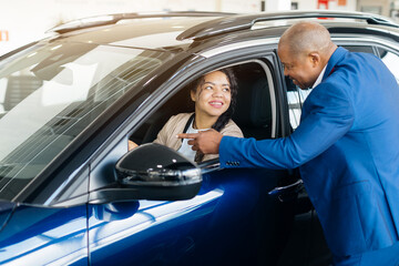 A young African American woman is interacting with a car dealer at a dealership. The man is...