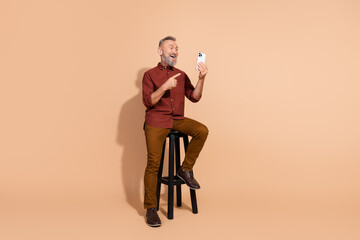 Senior man using smartphone while posing on a stool in a relaxed setting, showcasing modern technology and connectivity