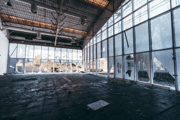 Interior of abandoned industrial building prepared for demolition with broken windows, shattered glass, damaged ceiling, debris on dirty floor, vandalism evidence, daylight scene