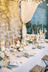 Long set table with bouquets of flowers and lit candles on the terrace of a stone building