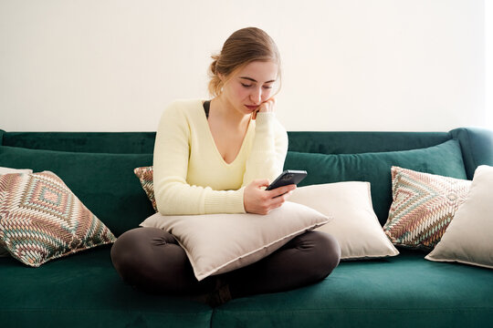 Boring young millennial woman using phone app, software, sitting on coach at home. Unhappy girl scrolling feed of social networks, spending time holding phone. Influence of social media, internet.