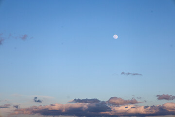 The photograph captures a clear blue sky with small cumulus clouds in the lower part of the frame. In the center of the sky, a pale moon is visible.