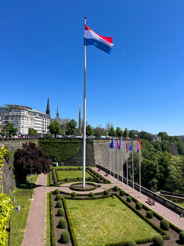 Garden next to the Plade De la Constitution, with the Luxembourg flag flying in the wind on a flagpole under a blue sky.