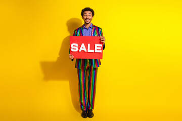 Happy young man in a colorful suit holding a red sale sign against a vivid yellow background,...