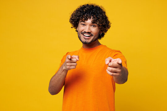 Young smiling fun happy Indian man he wear orange t-shirt casual clothes point index finger camera on you motivating encourage isolated on plain yellow background studio portrait. Lifestyle concept.