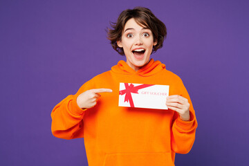 Young smiling happy woman wear orange hoody casual clothes hold point finger on gift certificate...