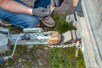 A man attaches a trailer to a car, securing safety chains to the trailer's hitch and drawbar