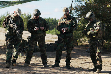 The military team of four stands in readiness, dressed in camouflage, inspecting weapons, unlocking pistols, adjusting rifles, and securing gear in holsters and belts.
