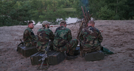 Fototapeta premium Hunters relax by the lake after a successful game hunt, grilling sausages over a bonfire, while their weapons are safely leaned against boxes behind them.