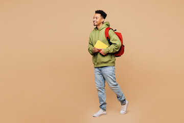 Full body side view young smart boy student wearing green casual clothes backpack bag hold books walking going isolated on plain pastel light beige background. High school university college concept. © ViDi Studio
