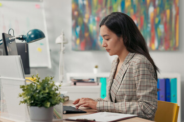 Attractive, experienced secretary of company is sitting in office in front of computer tapping...