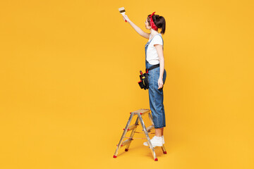 Full body young employee laborer handyman woman wear overalls belt stand on ladder paint isolated...