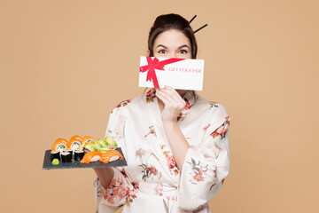 Young fun woman wear kimono clothes hold eat raw fresh sushi roll served on black cover mouth with plate gift certificate coupon voucher card isolated on plain beige background. Japanese food concept.