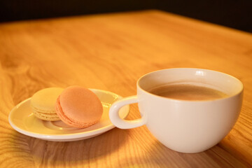 A warm beverage sits in a white cup next to a plate with two colorful macarons on a smooth wooden table, capturing a cozy afternoon moment at a cafe