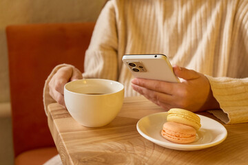 A person sits at a wooden table in a café, sipping coffee from a white cup and browsing on a smartphone. A plate with macarons sits nearby, creating a warm atmosphere