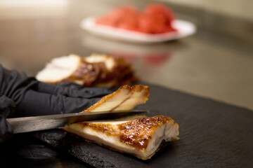 A chef with black gloves skillfully slices tender roasted meat on a slate board. Fresh ingredients can be seen in the background, enhancing the culinary atmosphere