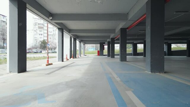 A drone captures an empty underground parking garage with concrete pillars and open sides. The industrial space is illuminated by natural light, creating a modern yet abandoned atmosphere