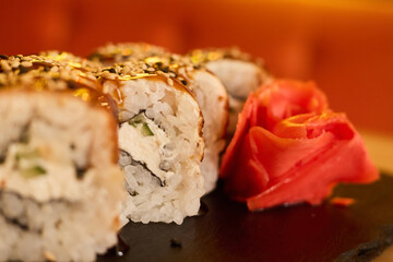 Fresh sushi rolls are artfully arranged on a black slate plate, garnished with a rose and sesame seeds. The warm ambiance enhances the dining experience