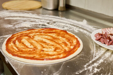 Fresh pizza dough spreads on a stainless steel surface, smeared with tomato sauce. Nearby, slices of meat await to be added, showcasing a busy kitchen atmosphere