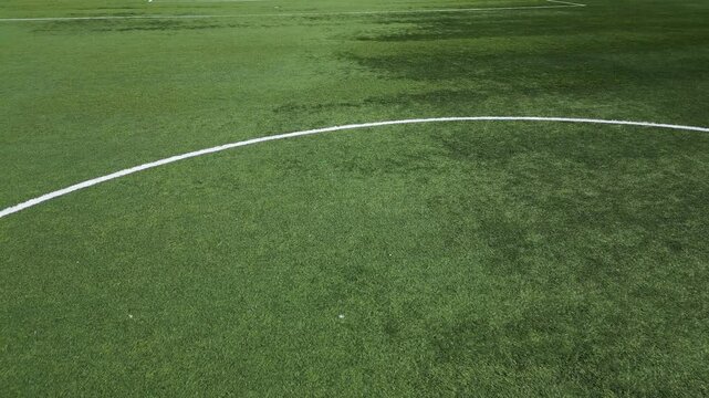 Low-altitude drone shot of an empty soccer field with artificial green grass, white boundary lines, and a goal net. The sports ground is well-maintained, creating a clean and structured composition