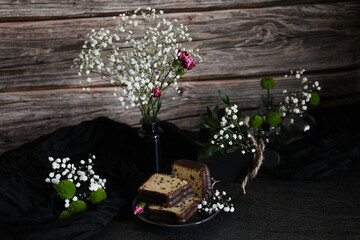 composition with gypsophila flowers in vases, pieces of cupcake on a plate and dark drapery against...