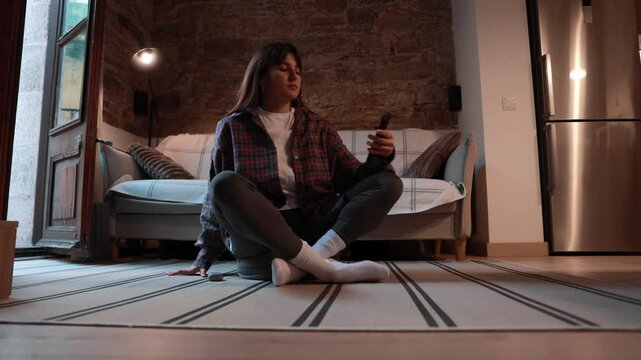 A young White woman wearing a plaid shirt, leggings, and socks, is sitting cross-legged on the floor, using her smartphone and wearing earphones in a modern living room.