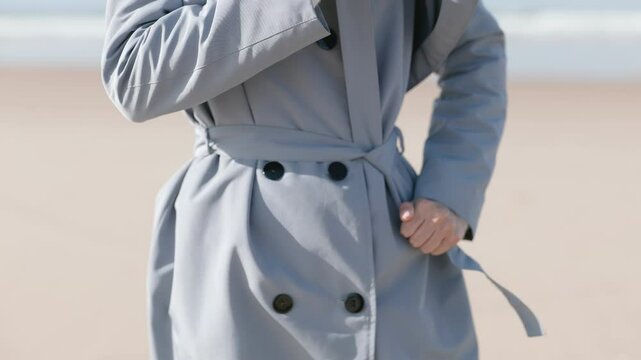 Close-up of a woman tying the belt of her stylish coat on a bright day at the beach, embodying casual elegance and carefree leisure.
