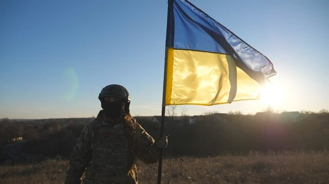 Young girl in camouflage uniform holds a waving flag of Ukraine against sunset. Female military medic of ukrainian army lifting blue-yellow banner on the field. Victory against russian aggression