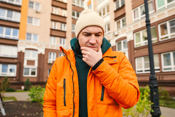 Man in Orange Jacket and Beige Beanie Thinks While Standing Outside an Apartment Building on a Cool...