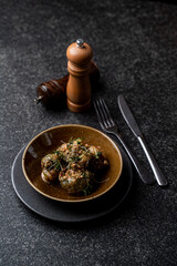 A beautifully arranged plate of lamb meatballs sits on a textured black surface, accompanied by silver cutlery and a wooden pepper grinder. Fresh herbs enhance the dish's appeal
