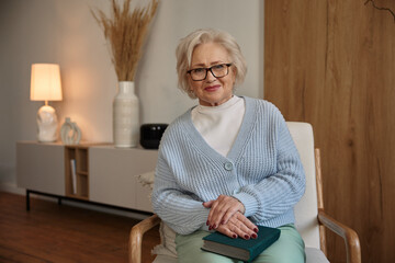 Attractive elderly woman wearing eyeglasses with book sitting on chair