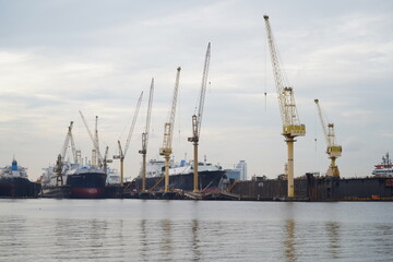Fototapeta premium Towering cranes operate at a busy shipping port, lifting containers with precision amid cargo ships, warehouses, and industrial structures.