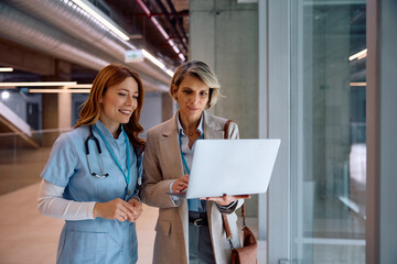 Businesswoman and nurse using laptop in hospital hallway.