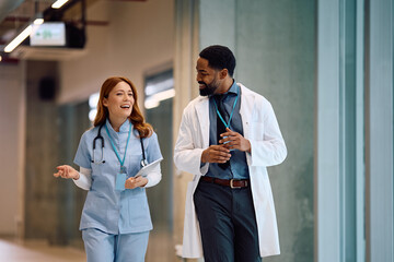 Happy nurse and black doctor talking while walking through hospital hallway.