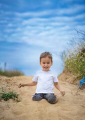 Child playing happily in sandy area. A young boy is sitting in soft sand, smiling brightly with a clear blue sky above. Surrounding greenery adds charm.