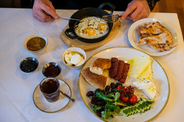 Turkish or Mediterranean style breakfast. Turkish breakfast. Variety of fresh vegetables, olives, cheeses, jams and spreads on small plates on white tablecloth covered table. Unrecognisable man hands