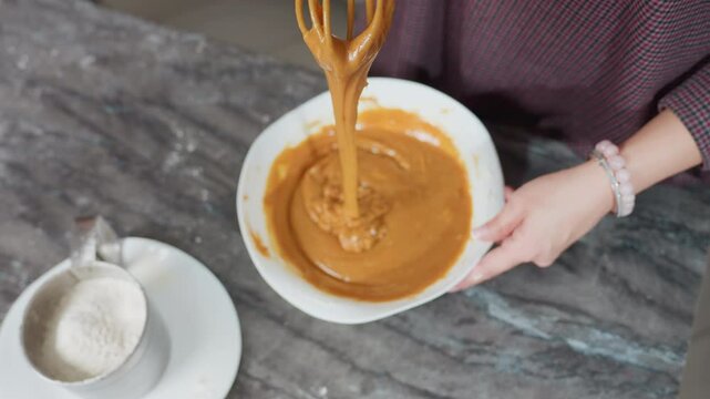 Top-down view of cook holding whisk with thick caramel-colored pap stretching as it drips back into white bowl, flour-dusted countertop and sifter nearby