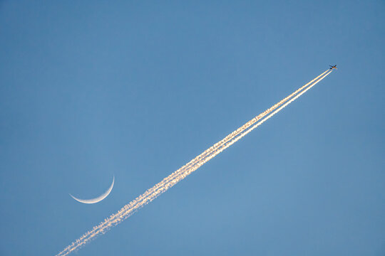 Airplane crossing sky near crescent moon, leaving vapor trail