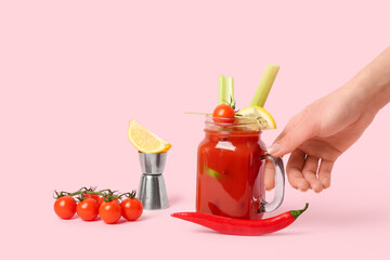 Female hand with mason jar of bloody mary cocktail and ingredients on pink background