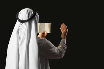 Young Muslim man with Koran praying on black background, back view. Ramadan celebration