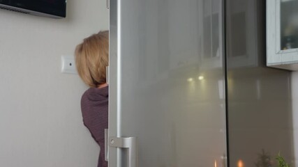 woman opens fridge to retrieve item, sleek door reflects kitchen background with warm lighting, wall socket visible near appliance, modern interior design with tiled walls
