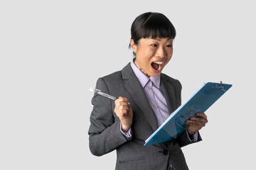 Photo of gorgeous Asian secretary woman with black hair, excitedly looking at the clipboard while writing down notes in the office, focused and engaged in her work.