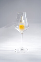 Cosmetic dropper bottle with yellow serum in a wine glass on white background. Aesthetic concept