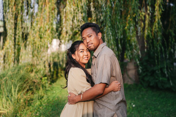 A loving Asian couple sharing a warm embrace in a lush green park, surrounded by cascading willow branches. The woman smiles softly, while the man looks confidently at the camera, reflecting love