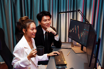 Smiling businesswoman in white suit looking on dynamic stock exchange statistic on pc screens holding coffee cup, comparing data sitting by coworker searching folder at night neon office. Infobahn.