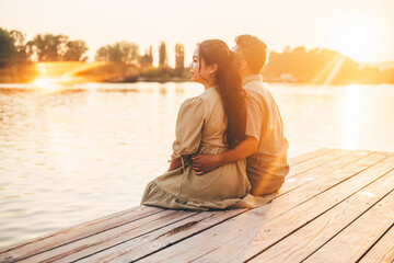 Fototapeta premium A loving couple sits closely together on a wooden pier, gazing at the serene lake as the golden sun sets on the horizon. The warm sunlight bathes them in a soft glow, creating a peaceful and intimate 