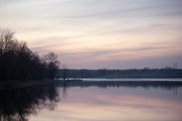 Fototapeta premium Golden hues blend with soft pastels as the sun sets over a serene lake. The calm waters mirror the silhouette of trees, creating a peaceful atmosphere at dusk