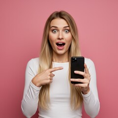 Fototapeta premium Surprised blond woman points at her smartphone with an excited expression against a pink background.