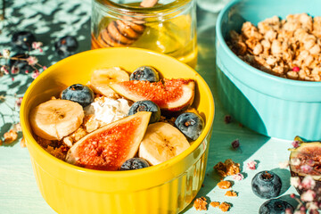 Granola with fresh banana, figs and blueberries on a light table with honey and flowers in a yellow and blue bowl. The concept of a delicious and healthy breakfast, a good start to the day